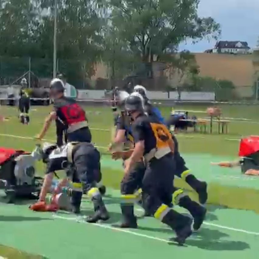 A group of people dressed in safety gear are running on a field. Some are tending to a person lying on the ground. Trees and a house are in the background.