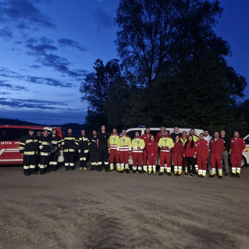 A group of firefighters dressed in red and yellow uniforms poses for a photo in the evening. They stand in front of red vehicles on a dirt road.