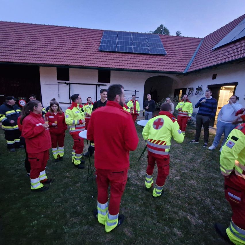 A group of firefighters in red and yellow uniforms are gathered in front of a house with solar panels on the roof. They are standing on grass.