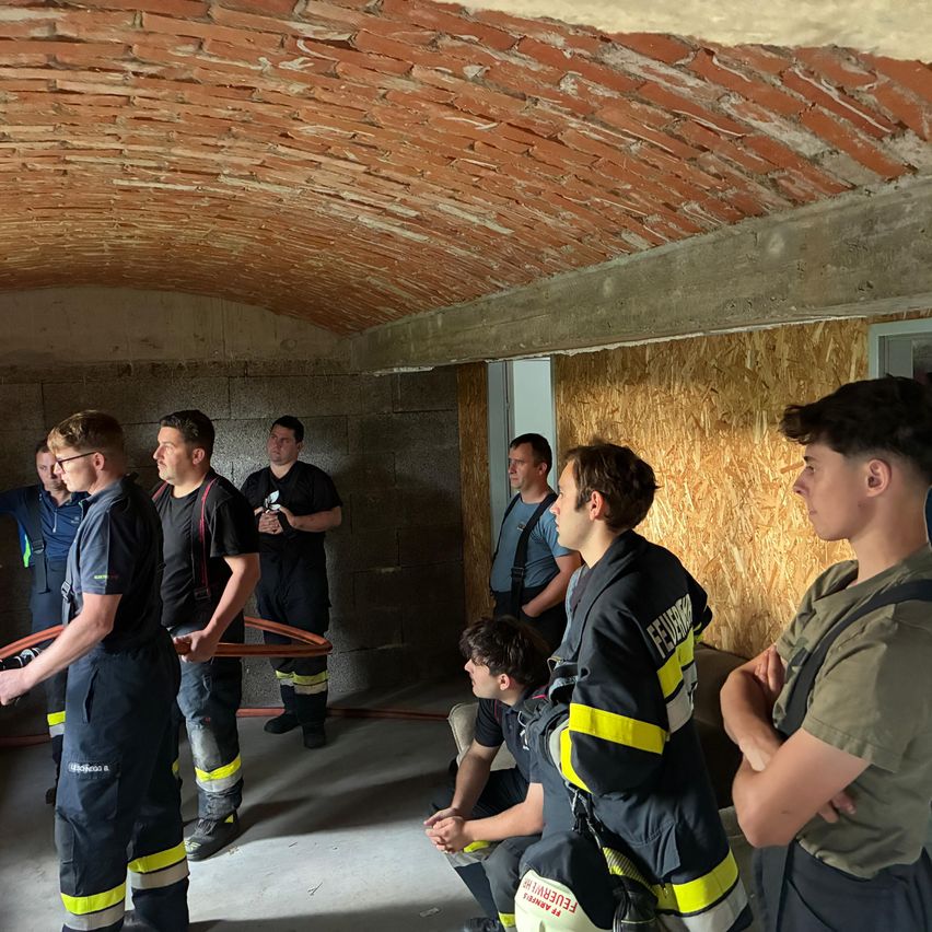Eine Gruppe von Feuerwehrleuten versammelt sich in einem Raum mit einer Ziegelbogendecke. Sie tragen schwarze Uniformen mit gelben und weißen Streifen. Einige stehen, andere sitzen. Einer hält eine Feuerwehrschlauch.