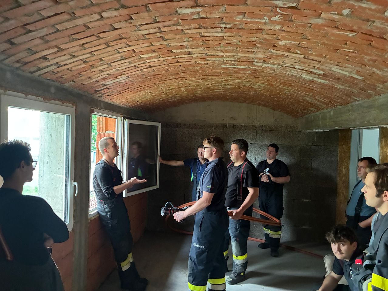 Fünf Männer in Uniformen stehen in einem unfertigen Raum, einer hält eine Feuerwehrhose, ein anderer gestikuliert. Ein Fenster spiegelt einen Mann wider. Die Decke ist mit Ziegeln gewölbt.