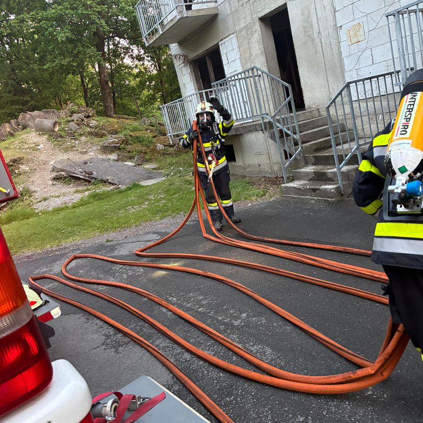 Feuerwehrleute sind dabei, einen Feuerwehrschlauch vor einem Gebäude mit Treppen und Balkon zu verlegen. Ein Feuerwehrmann hält den Schlauch, während ein anderer bereit steht.