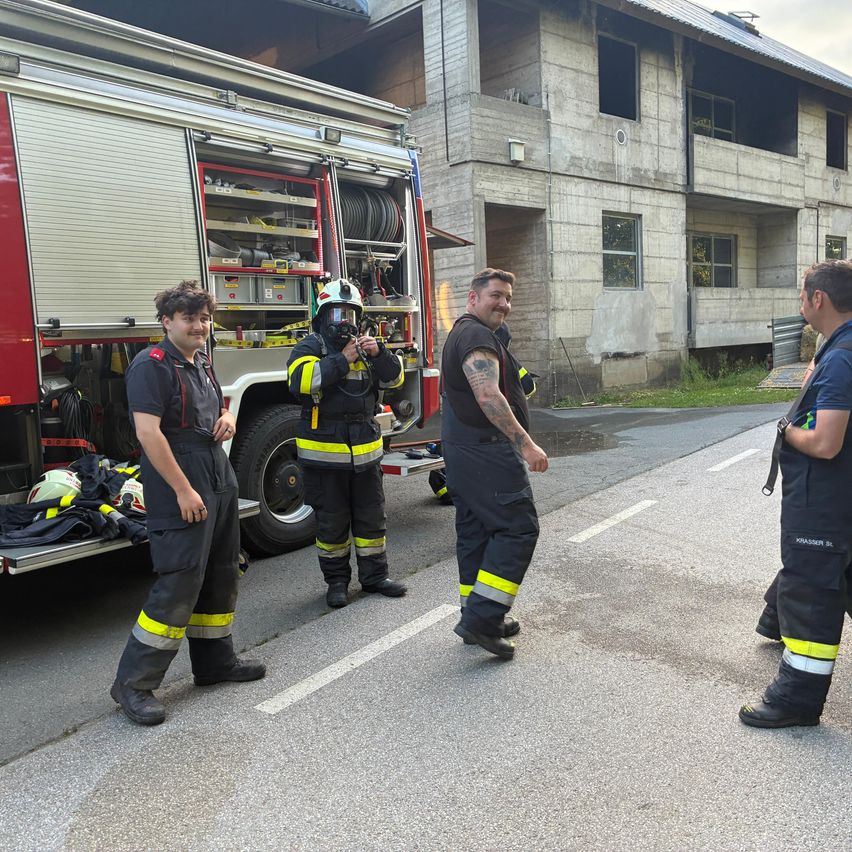 Feuerwehrleute in voller Ausrüstung stehen neben einem Feuerwehrwagen und besprechen ihren nächsten Schritt vor einem beschädigten Gebäude.