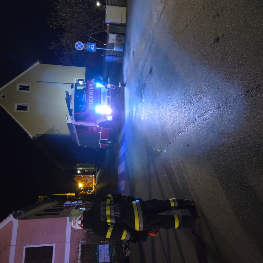 Ein Feuerwehrmann in Uniform steht nachts vor einem Gebäude, das von den Lichtern eines Feuerwehrwagens beleuchtet wird.