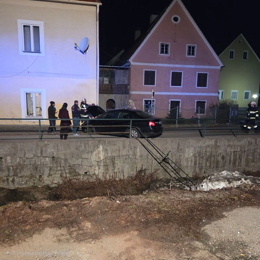 In der Nacht ist ein Auto in der Nähe eines Gebäudes geparkt, mit Feuerwehrleuten und Polizeibeamten. Das Auto hat einen kaputten Stoßfänger an der Betonbarriere.