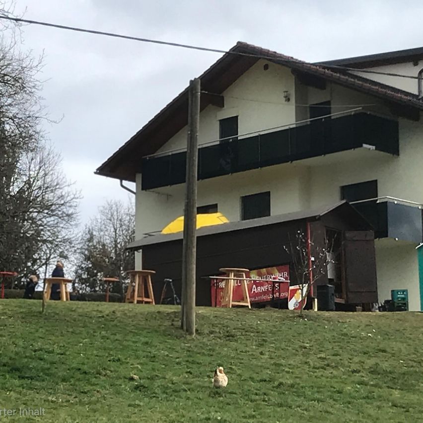 Ein Haus mit Balkon und gelbem Schirm davor. Vor dem Haus stehen einige Tische und Stühle. Rechts befindet sich ein kleines Gebäude mit einem roten Schild.