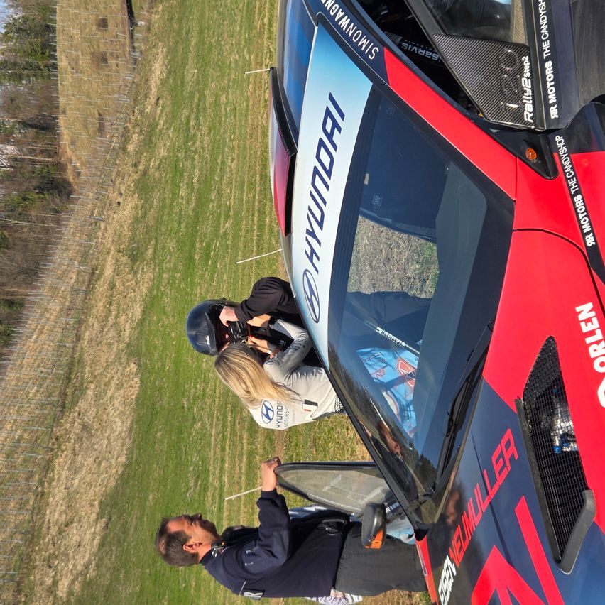 Ein roter Hyundai Rallye-Wagen mit dem Wort Hyundai an der Seite. Zwei Personen verstellen etwas am Auto, einer von ihnen hält eine Kamera.