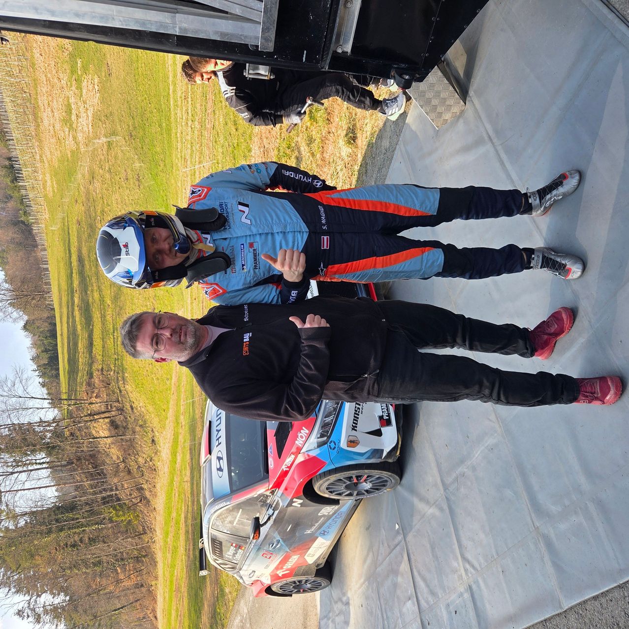 Drei Personen stehen neben einem Rennwagen, zwei in Rennausrüstung, einer in einem schwarzen Jackett. Dahinter befindet sich eine Rasenfläche und Bäume.