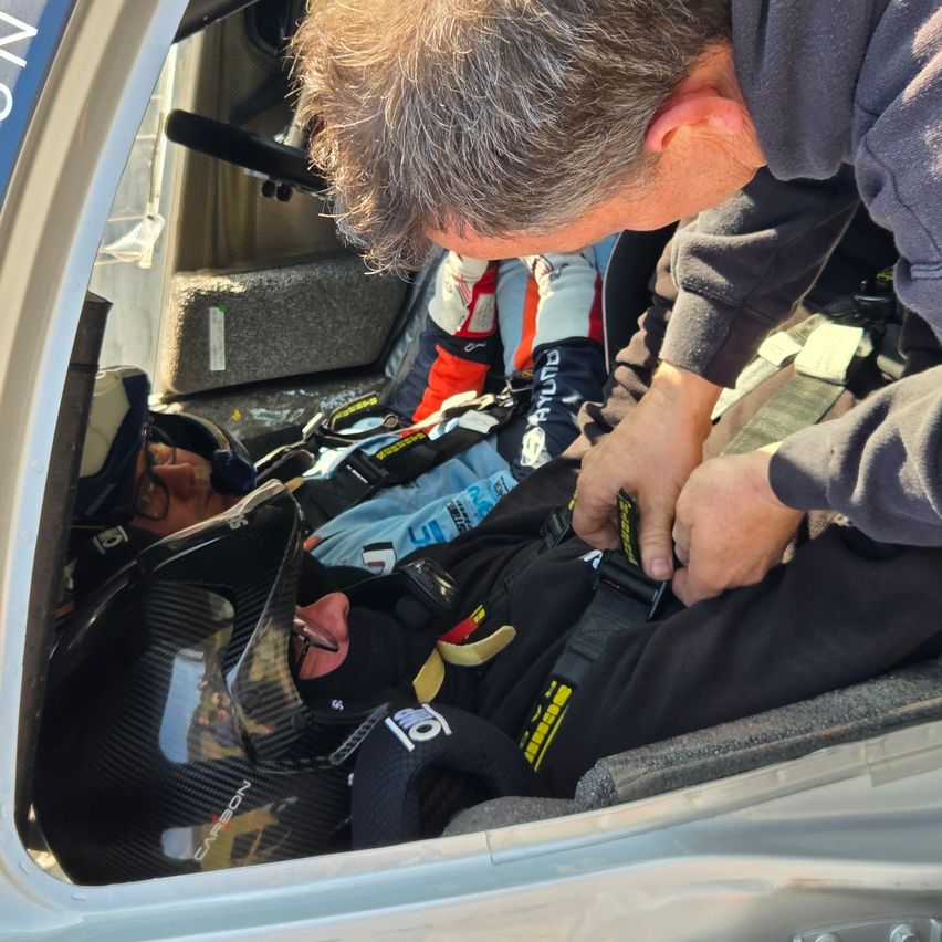 Eine Person in einem Hubschrauber hilft einer anderen Person, die einen Helm und eine Schutzbrille trägt, einen Sicherheitsgurt anzulegen.