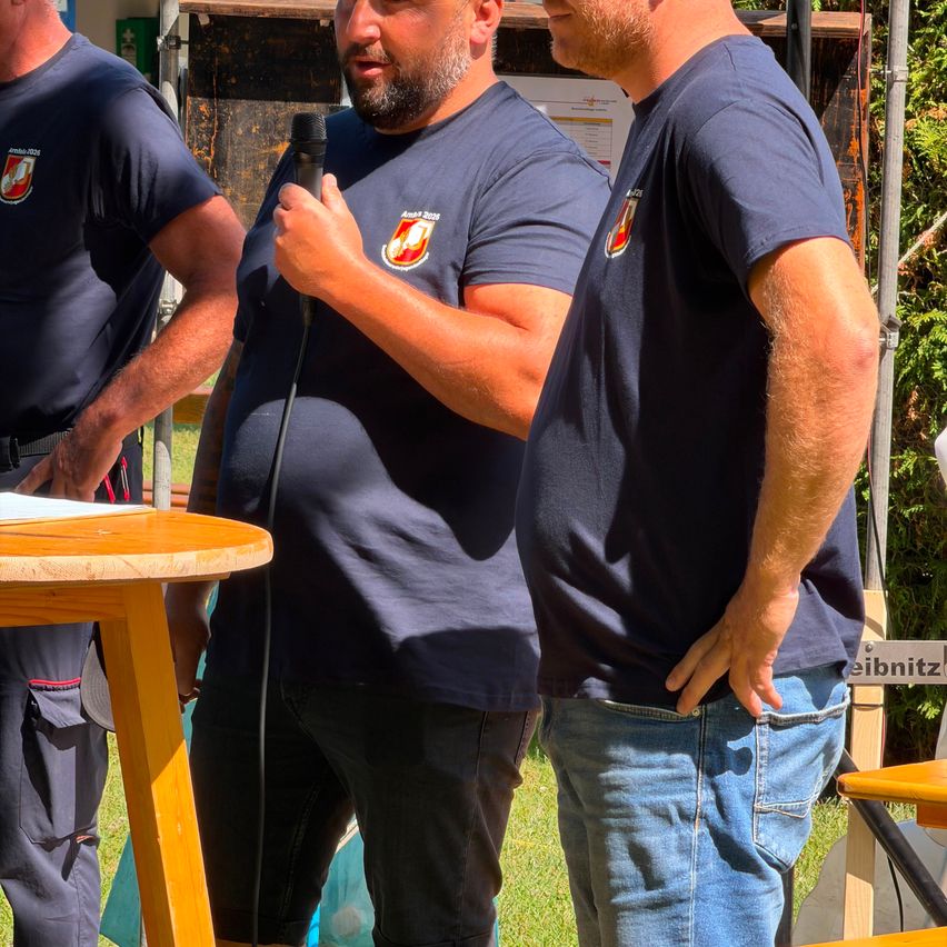 Drei Männer in blauen Uniformen stehen draußen. Einer hält ein Mikrofon, ein anderer hat ein Papier und der dritte hat die Hände auf den Hüften.