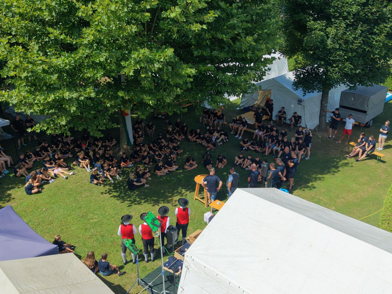 Eine Gruppe von Menschen in einheitlichen Kleidungsstücken versammelt sich im Freien. Einige sitzen auf dem Rasen, während andere stehen. Es gibt Zelte und einen Tisch in der Umgebung.