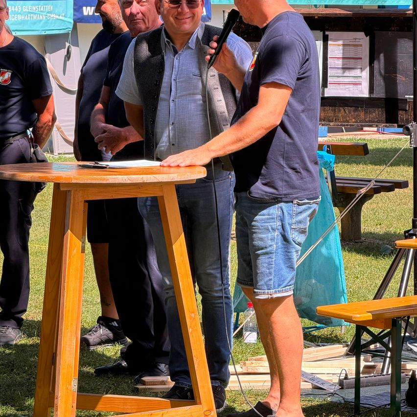 Eine Gruppe von Männern steht um einen Holztisch auf einem Rasen. Ein Mann hält ein Mikrofon und scheint zu sprechen. Hinter ihnen ist eine Struktur mit einem Banner und einer Bank zu sehen.