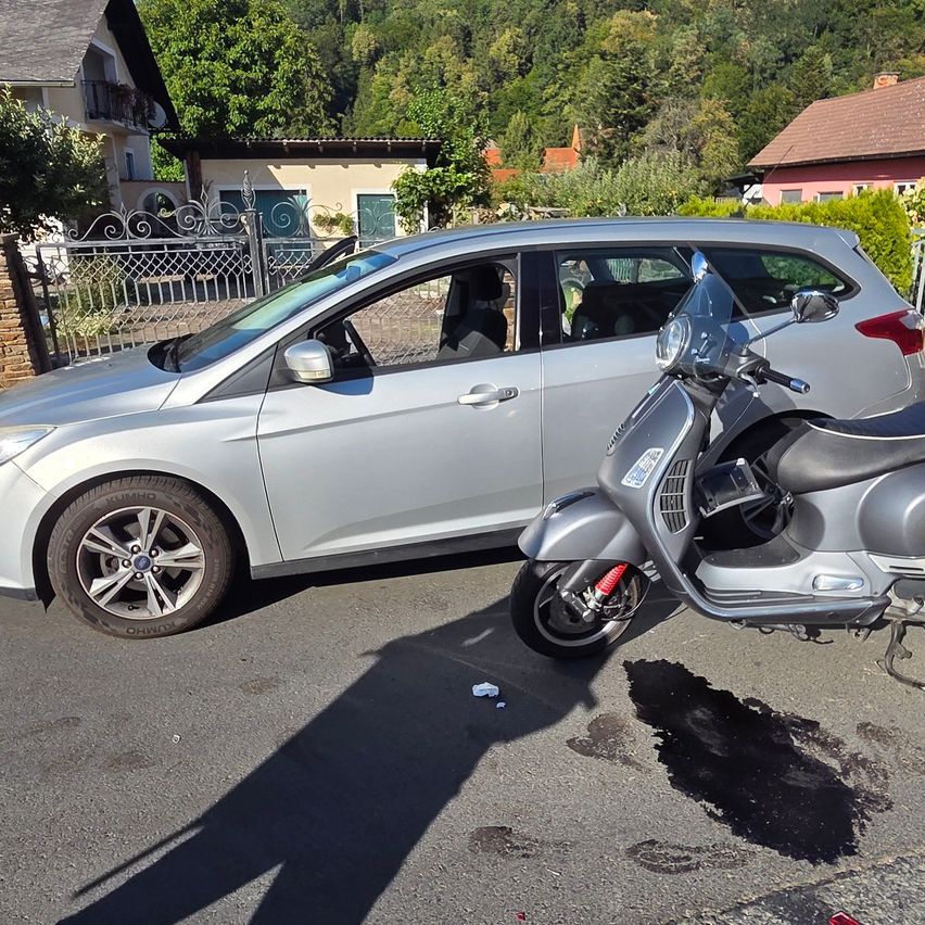 A silver car and a silver scooter are parked on the side of the road with a small oil spill on the ground.