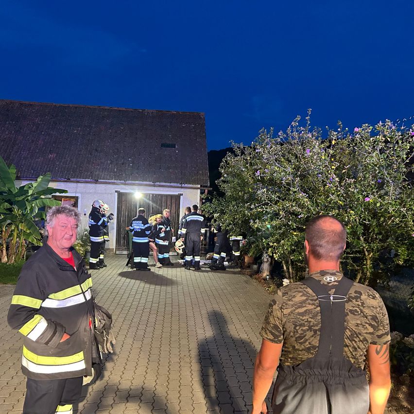 Eine Gruppe von Feuerwehrleuten versammelt sich nachts vor einem Haus, wobei ein Mann in einer reflektierenden Weste zusieht.