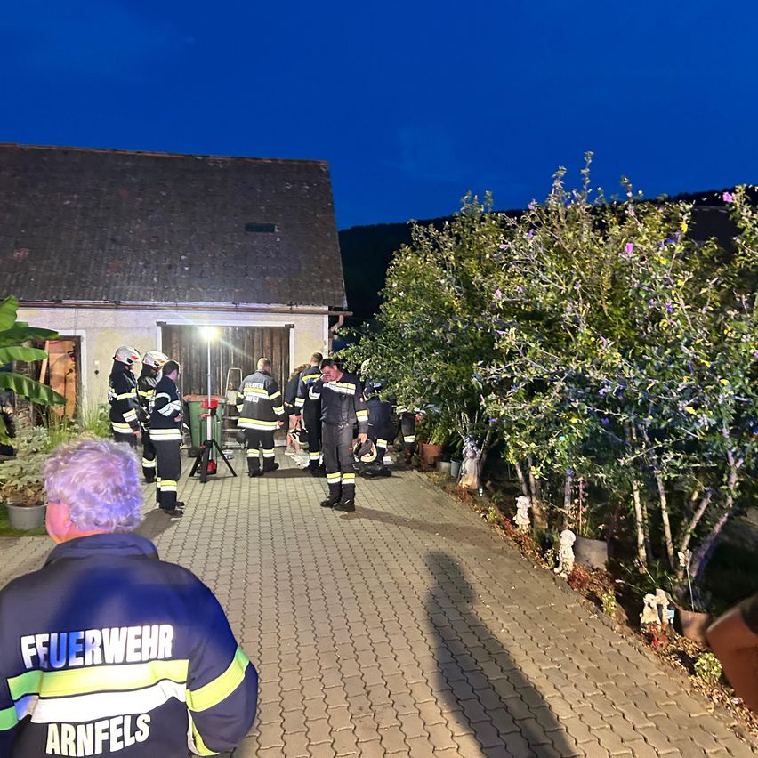 Eine Gruppe Feuerwehrleute in Uniform steht nachts vor einem Haus, eine Person hält einen Helm.