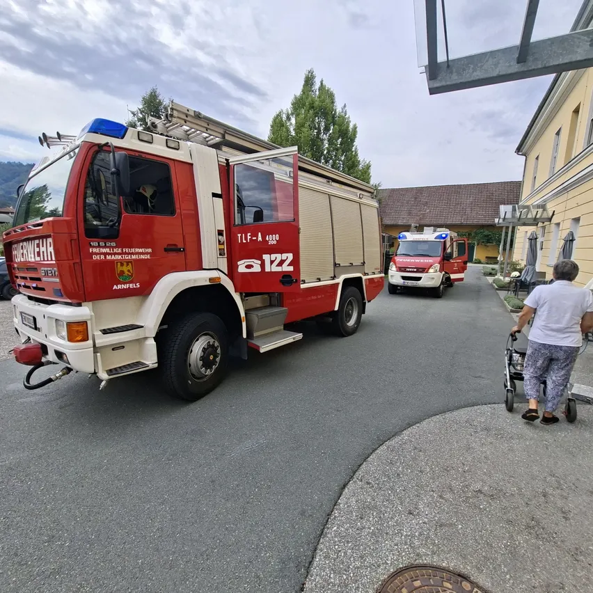 Ein Feuerwehrauto ist am Straßenrand geparkt, mit einem weiteren Feuerwehrauto im Hintergrund. Im Vordergrund schiebt eine Person einen Rollstuhl.