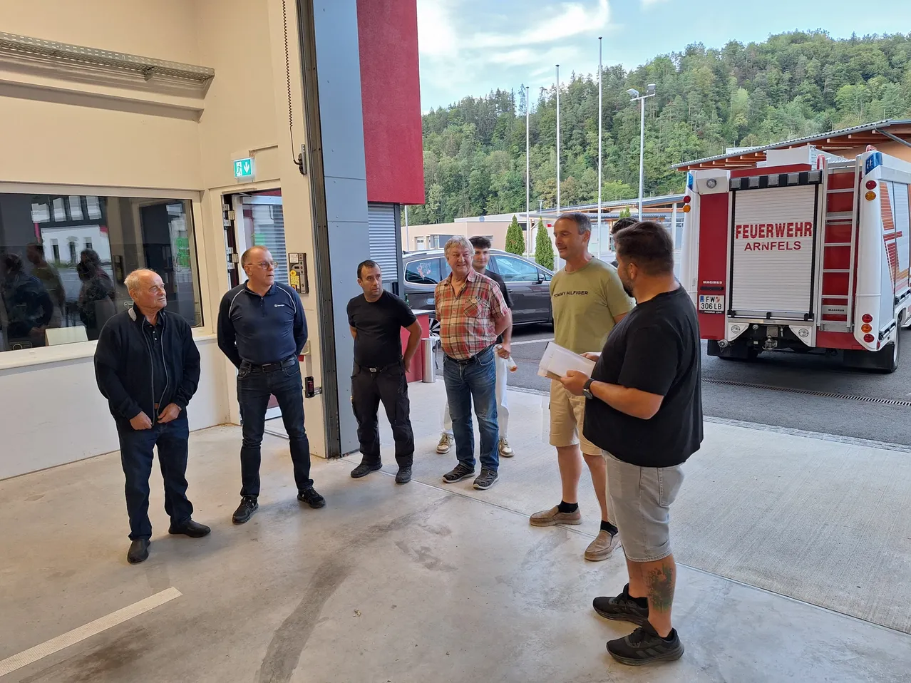 Sechs Männer stehen in einer Garage, zwei von ihnen halten Papiere. Ein Feuerwehrauto ist rechts geparkt.