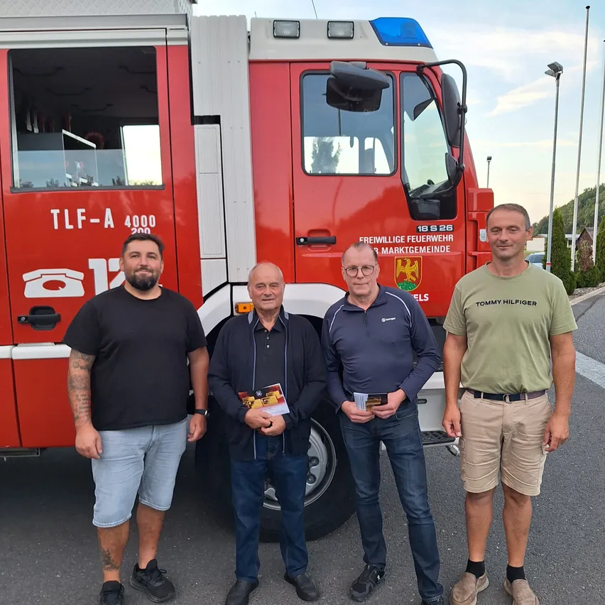 Vier Männer stehen vor einem roten Feuerwehrauto mit der Aufschrift TLF-A 4000. Sie halten Flugblätter in den Händen und lächeln für die Kamera.