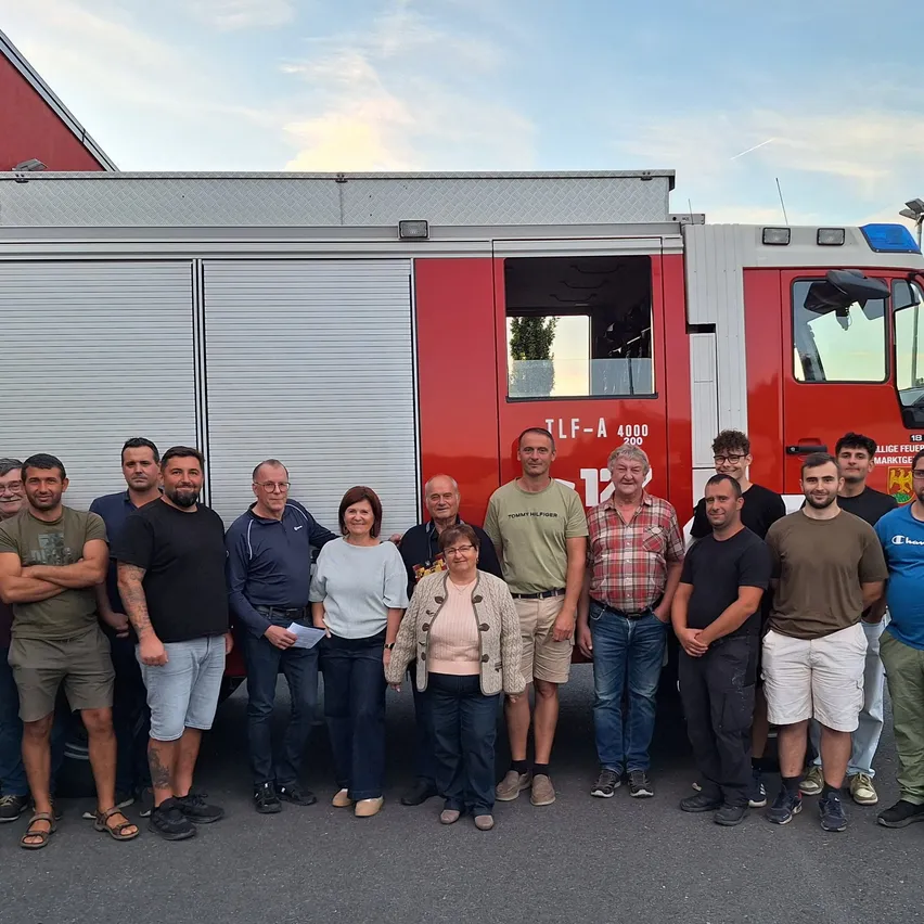 Eine Gruppe von Feuerwehrleuten und Freiwilligen posiert vor einem roten Feuerwehrauto. Sie alle lächeln und scheinen für ein Foto zu posieren.