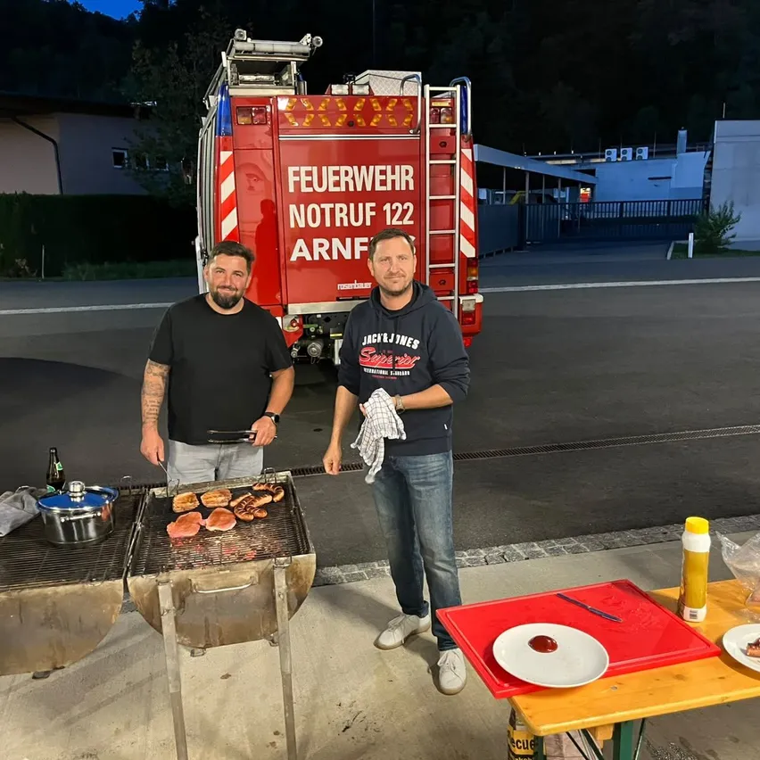 Zwei Männer stehen vor einem Feuerwehrauto und grillen Fleisch auf einem Grill. Einer hält eine Pfanne, der andere ein Handtuch. Ein Tisch mit Tellern und Gewürzen ist in der Nähe.