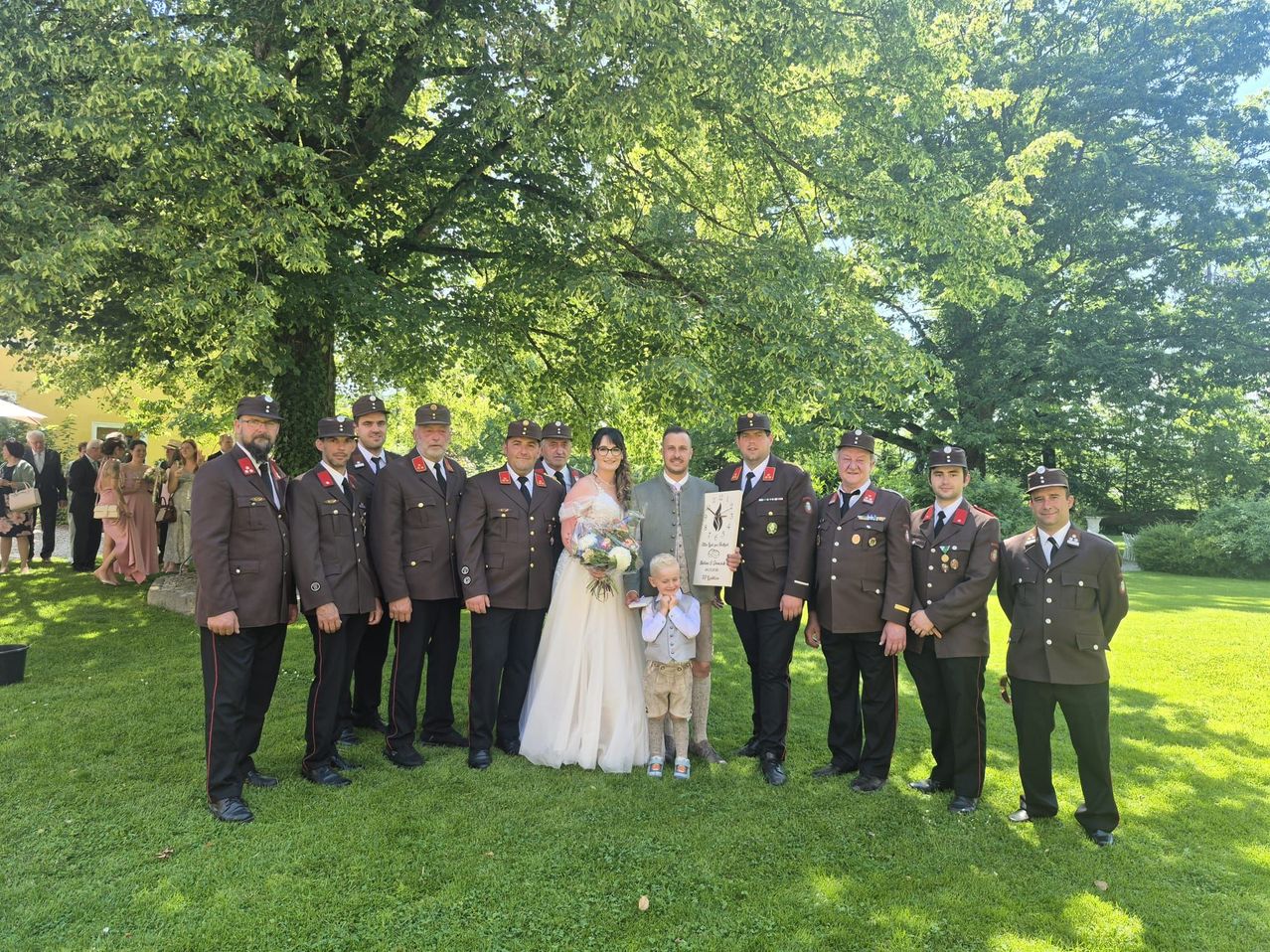 A wedding party with military officers and a family in uniforms poses in a park with green trees and grass.
