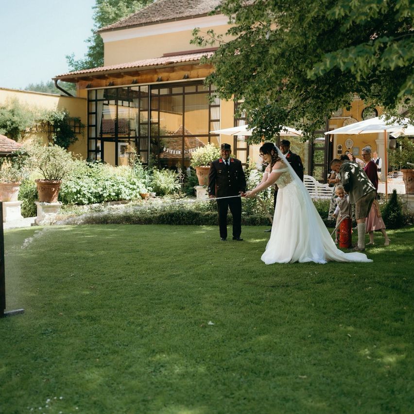 A bride in a wedding gown holds a sword in a garden with a uniformed man. Behind them are potted plants, a building, and people.