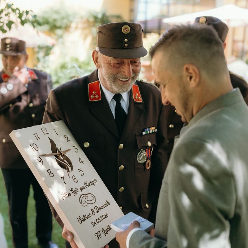 An older man in a military uniform presents a wooden clock to another man in a ceremony. The clock features the numbers 1-12 and the words 'Sabine & Dominik'.
