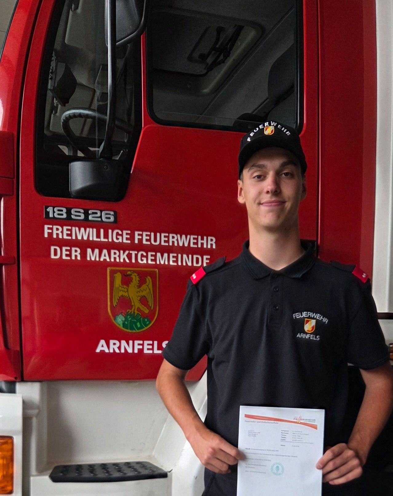 A young man in a black uniform stands in front of a red fire truck. He holds a certificate and smiles. The fire truck has the words Freiwillige Feuerwehr Der Marktgemeinde Arnfels on it.