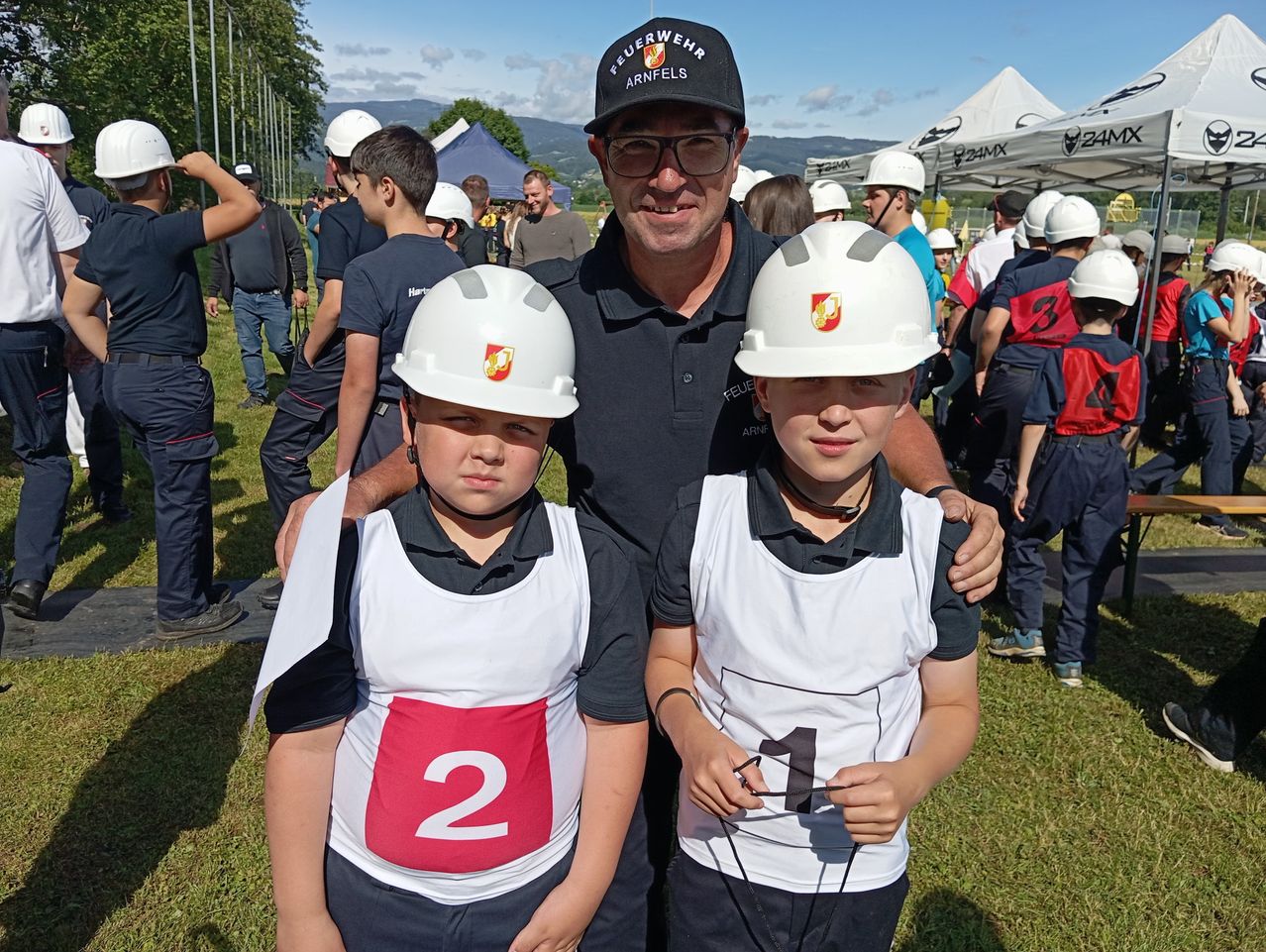 A smiling man with two young boys wearing helmets stands in a field. They are holding a number sign, possibly for a competition. There are tents and people in the background.