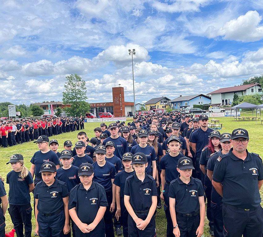 A large group of young people in uniform standing in a field. The group is organized into rows, and they are all looking forward. There are several adults in the group, and they are also in uniform. The area has several trees and houses in the background.