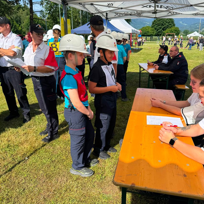 A group of children in safety gear stand in line while adults review documents under a tent. Nearby, a woman signs a paper.