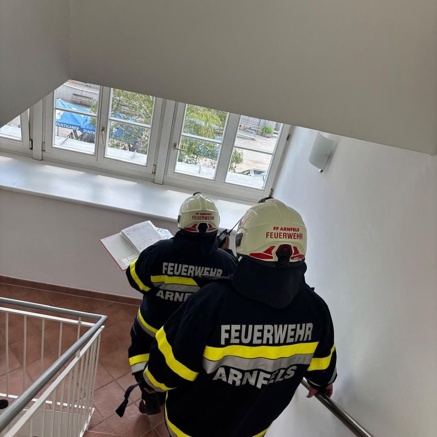 Zwei Feuerwehrleute in Uniformen mit der Aufschrift 'Feuerwehr' und 'Arnfels' steigen eine Treppe hinauf. Sie tragen Helme und tragen Ausrüstung. Ein Fenster mit Blick auf ein blaues Zelt und Bäume befindet sich hinter ihnen.
