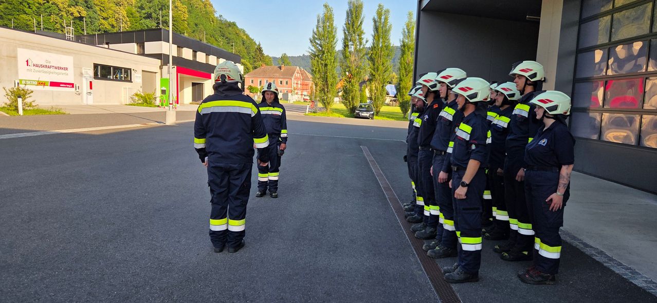 Feuerwehrleute in Uniform stehen in einer Reihe draußen mit einem geparkten Auto im Hintergrund.