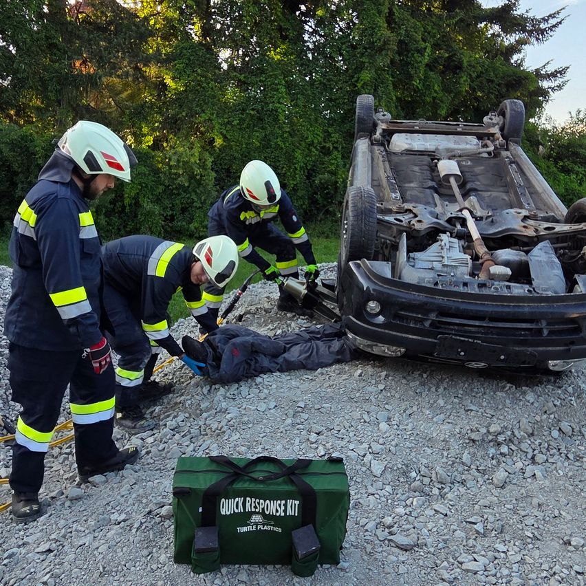 Notfallhelfer in Schutzkleidung arbeiten an einem umgekippten Auto. Eine Tasche mit der Aufschrift 'Quick Response Kit' befindet sich in der Nähe.