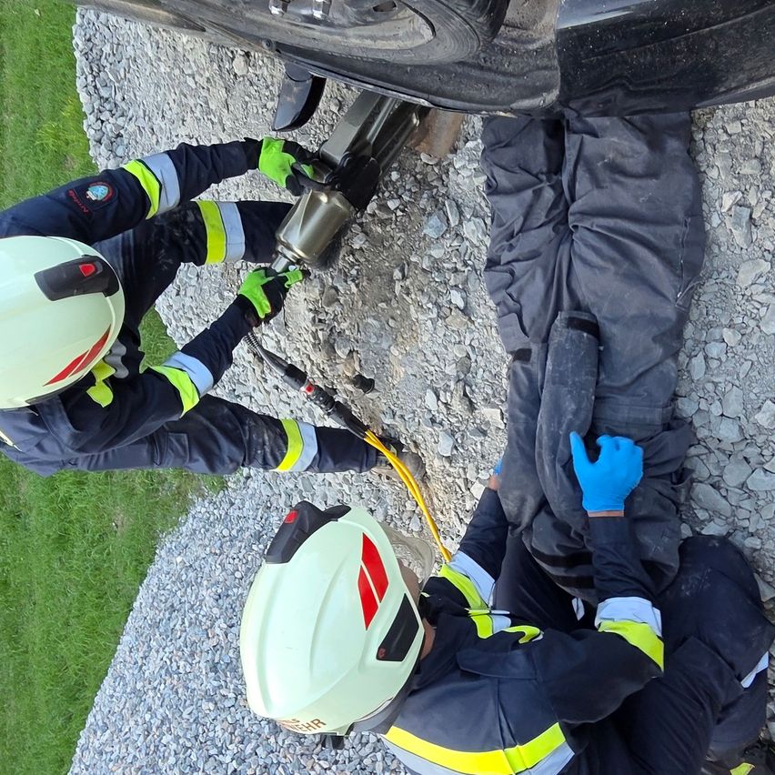 Zwei Personen in Schutzkleidung führen eine Rettungsaktion durch. Eine hält ein Werkzeug unter einem Auto, während die andere auf dem Boden liegt, beide tragen Helme und Handschuhe.