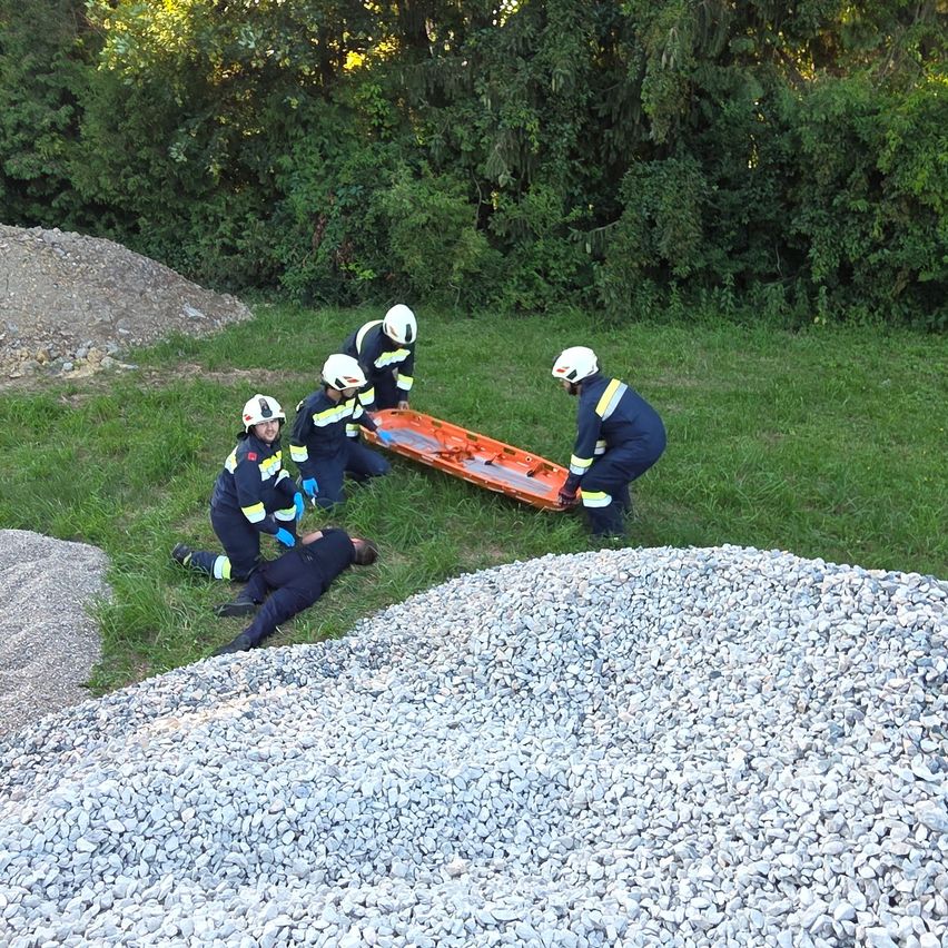 Fünf Rettungskräfte bereiten sich darauf vor, eine verletzte Person mit einer Trage in einem offenen Bereich mit Kies und Bäumen zu transportieren.