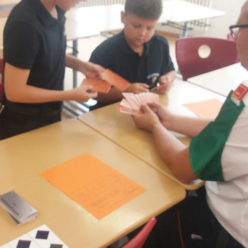 Zwei Jungen in schwarzen Hemden spielen ein Kartenspiel, wobei einer ein Deck hält und der andere eine Karte betrachtet. Ein Erwachsener in einem grün-weißen Hemd beobachtet sie.
