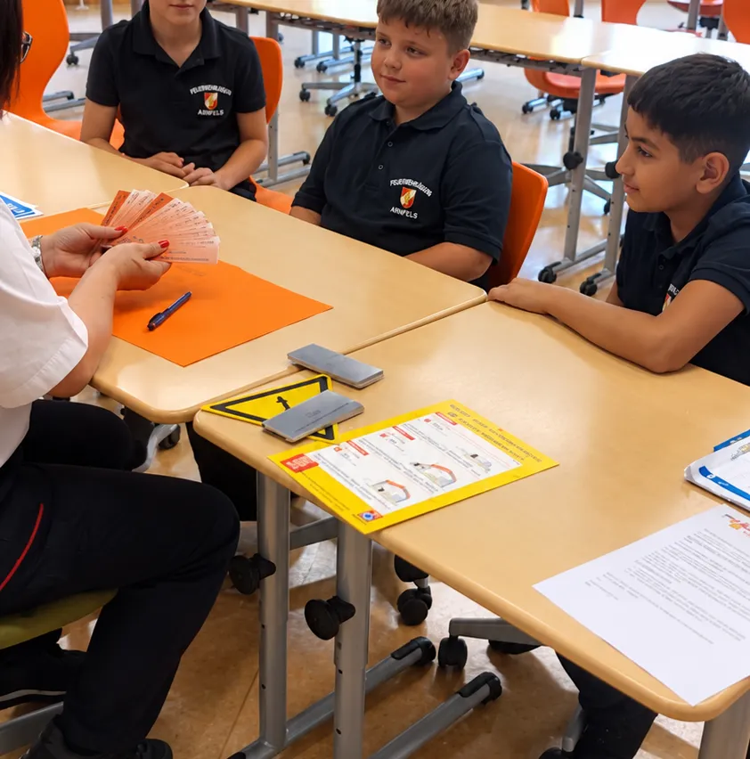 Ein Klassenzimmer mit Schülern an Tischen, ein Schüler hält Stift und Papier, ein anderer Schüler schaut auf eine Karte.