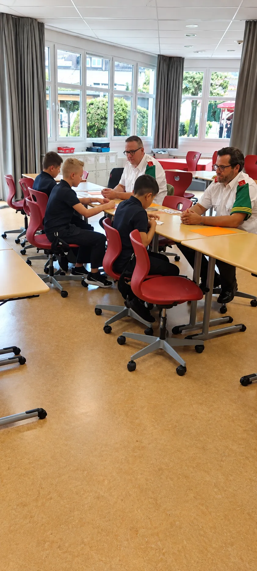 Vier Jungen sitzen um einen Tisch herum mit einem Mann in Uniform. Sie scheinen ein Kartenspiel zu spielen. Der Raum ist gut beleuchtet und hat ein modernes Design.