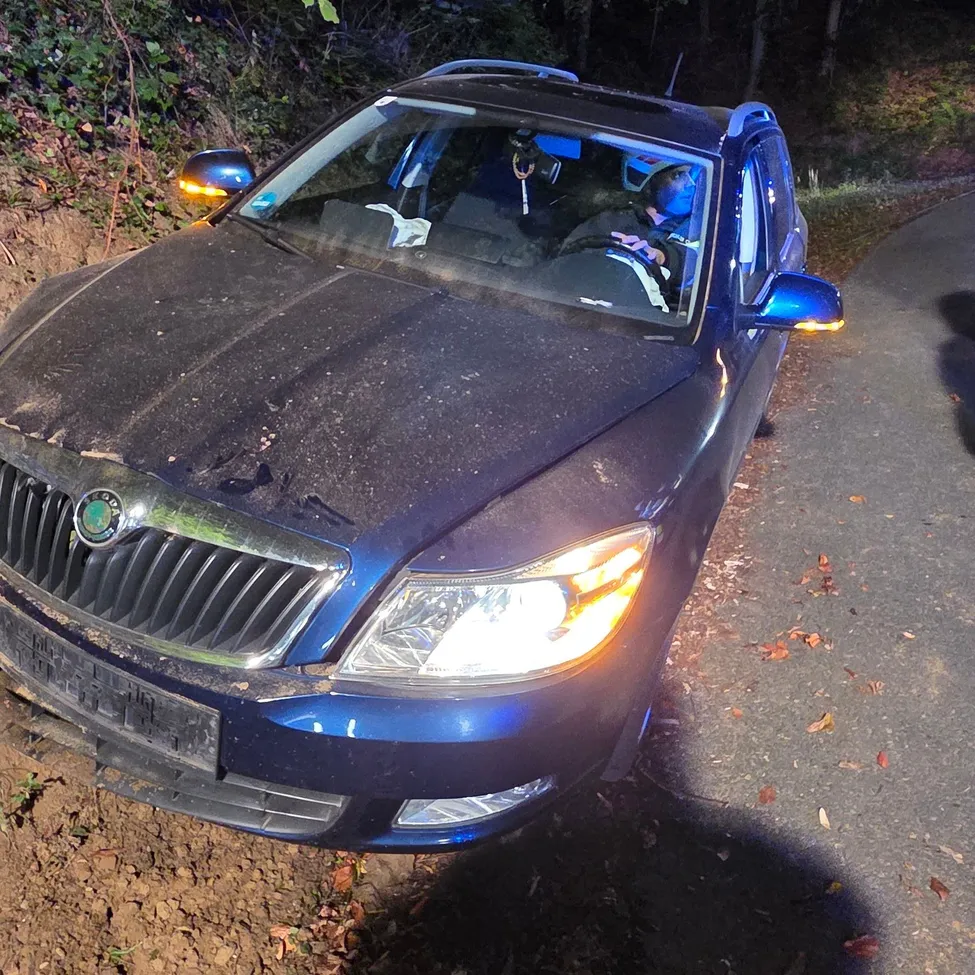 Ein blaues Auto ist nachts auf einer unbefestigten Straße geparkt. Das Auto ist zur Seite geneigt und die Scheinwerfer sind eingeschaltet.