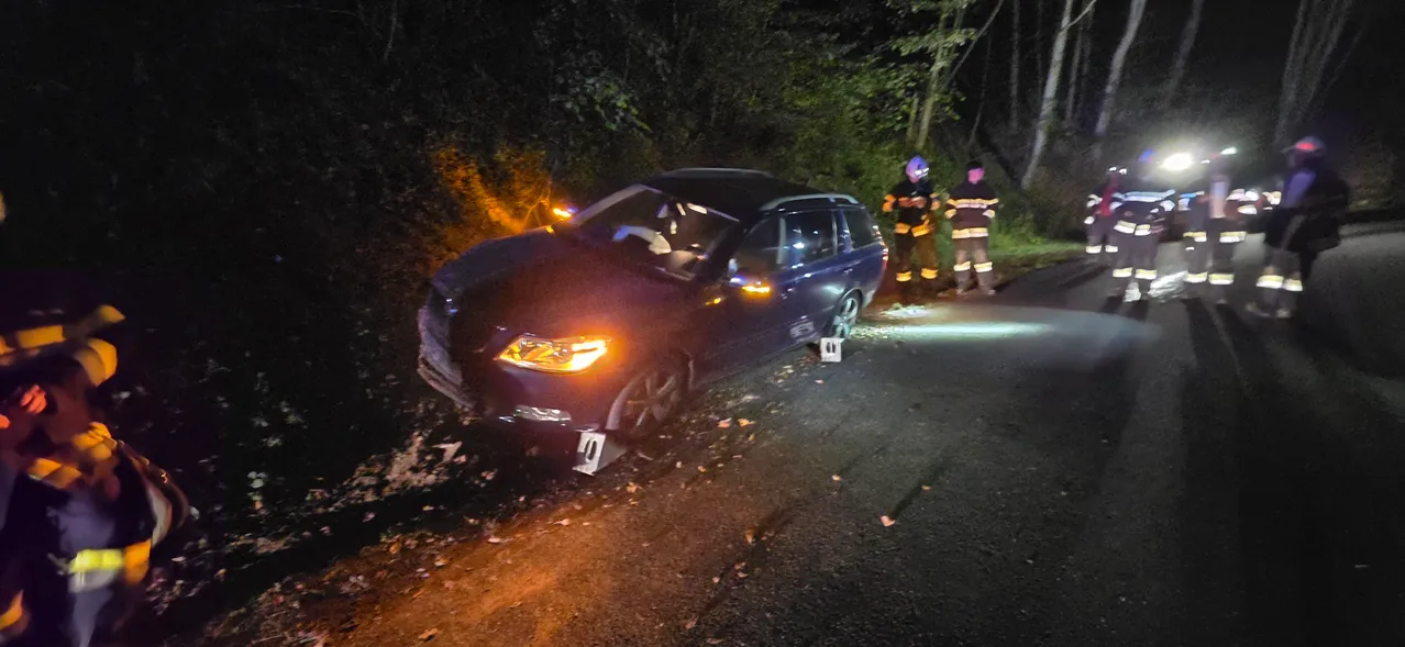 Eine dunkle Nachtszene mit einem beschädigten Auto am Straßenrand. Mehrere Personen in Sicherheitsausrüstung stehen um das Auto herum, möglicherweise Feuerwehrleute.
