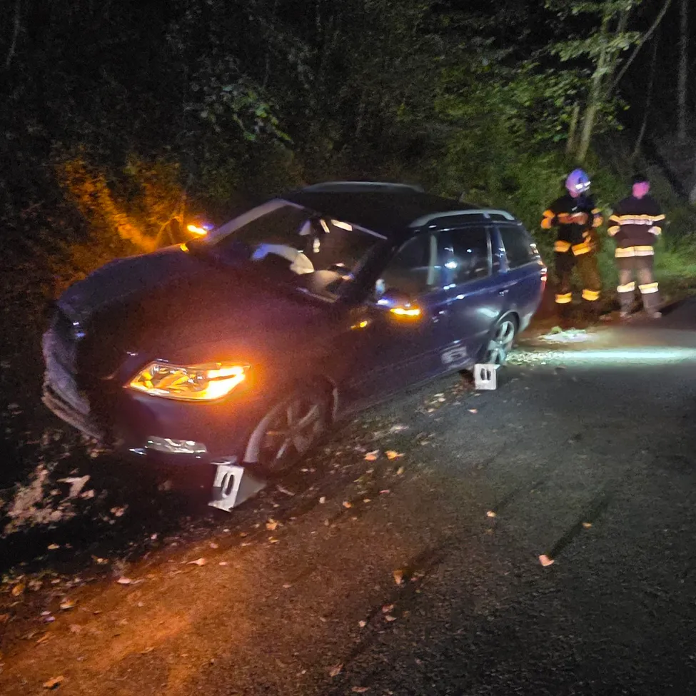 Eine dunkle Nachtszene mit einem beschädigten Auto am Straßenrand. Mehrere Personen in Sicherheitsausrüstung stehen um das Auto herum, möglicherweise Feuerwehrleute.