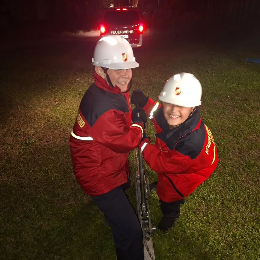Zwei Personen in Feuerwehruniformen stehen nachts auf einer Leiter. Die Person auf der linken Seite hält die Leiter, während die Person auf der rechten Seite lächelt.