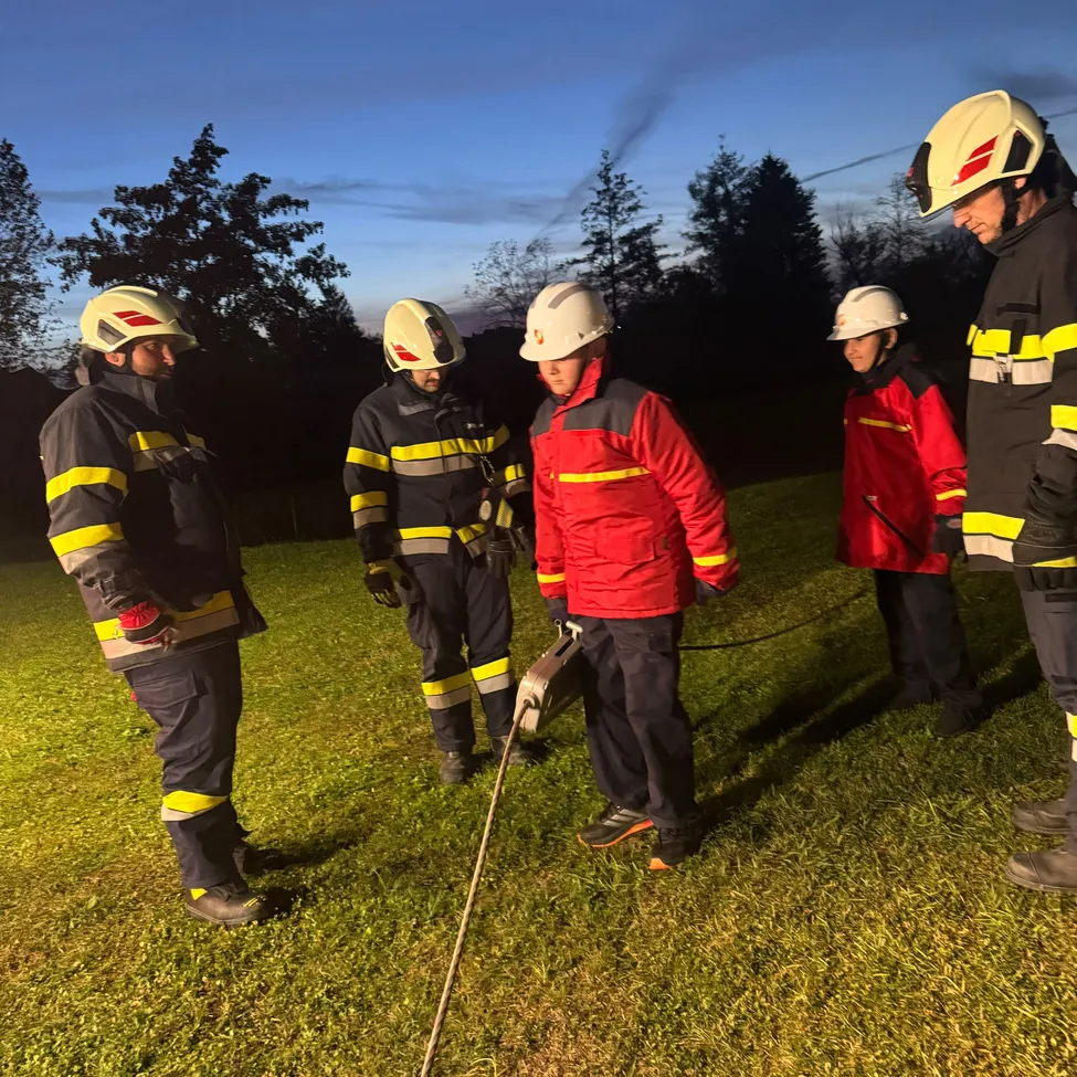 Fünf Feuerwehrleute in voller Ausrüstung stehen auf einem Grasfeld bei Dämmerung. Sie halten ein Seil und scheinen etwas zu besprechen.
