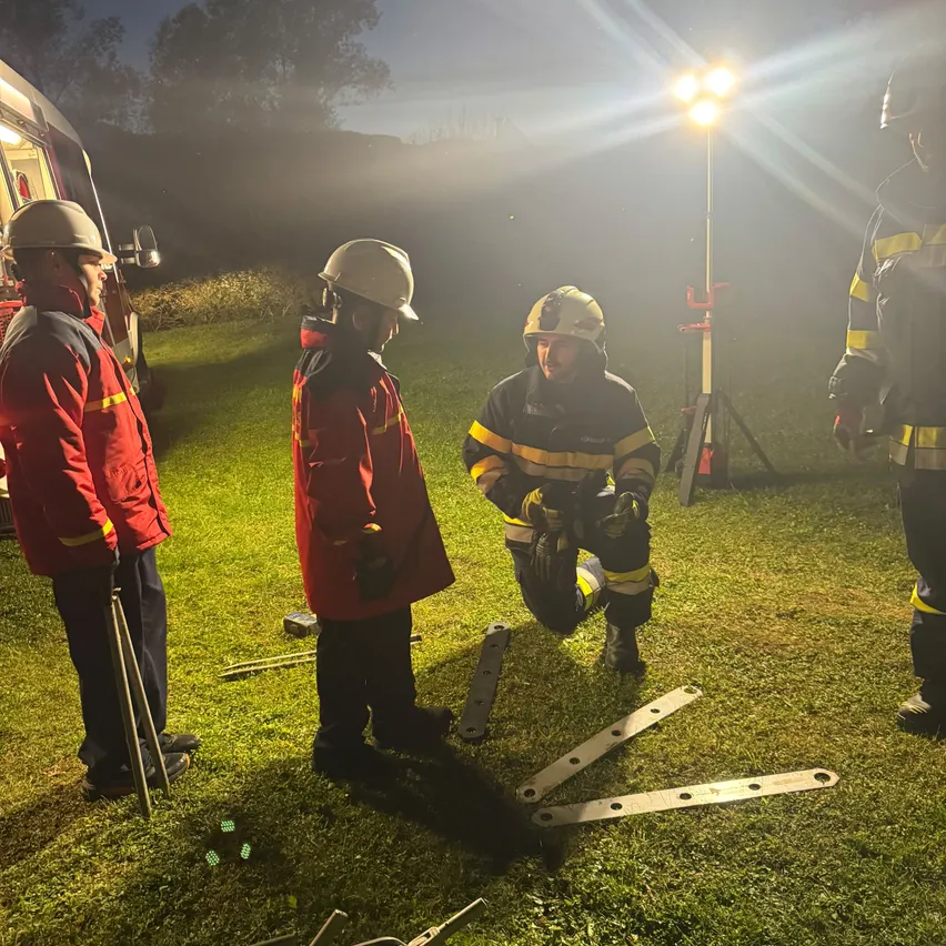 Fünf Feuerwehrleute in roten Uniformen versammeln sich nachts auf einem Rasenfeld. Einer kniet und scheint etwas zu reparieren, während die anderen stehen und zusehen.