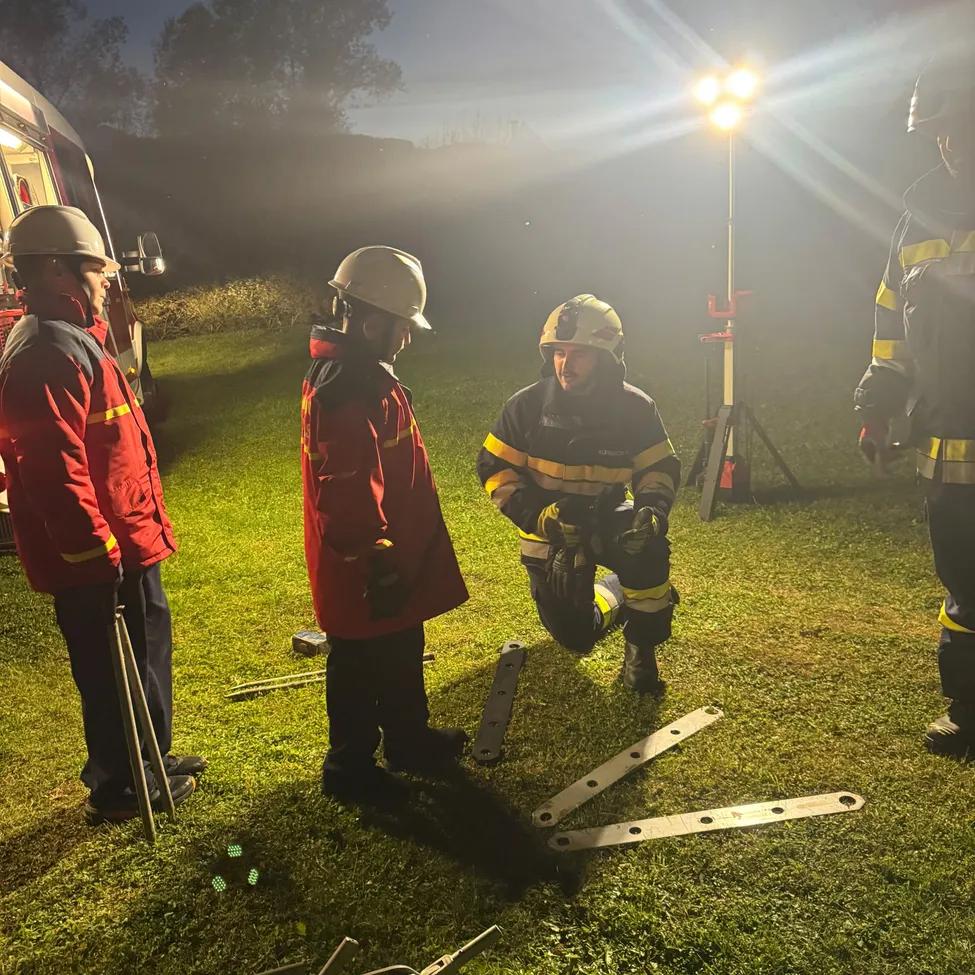 Fünf Feuerwehrleute in roten Uniformen versammeln sich nachts auf einem Rasenfeld. Einer kniet und scheint etwas zu reparieren, während die anderen stehen und zusehen.