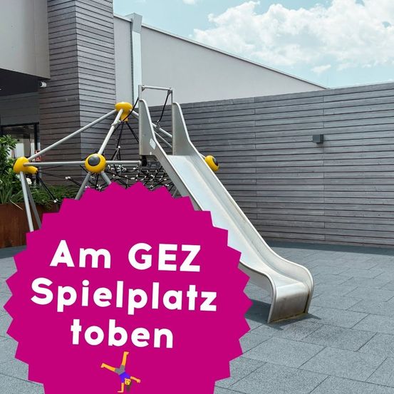 Ein Spielplatz im Freien mit einer Rutsche und Klettergerüst, umgeben von einem Gebäude und Grünflächen. Die Rutsche ist silber, und das Klettergerüst ist gelb. Der Text 'Am GEZ Spielplatz toben' steht in Weiß auf einem rosa Hintergrund.
