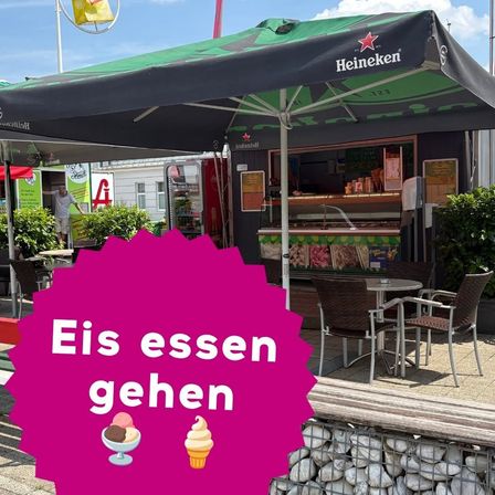 Ein Eisstand mit einem großen grünen und schwarzen Regenschirm, der verschiedene Eissorten anbietet, und einer Person, die hinter der Theke steht.