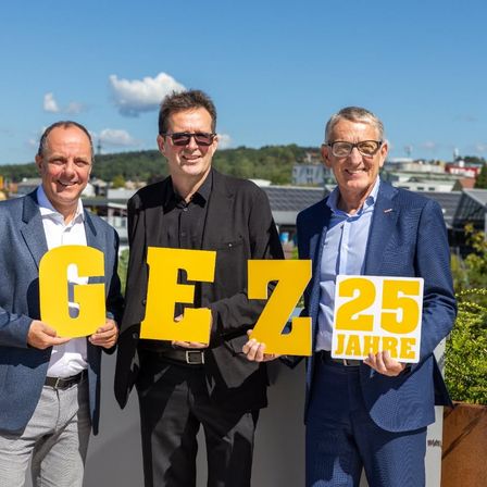 Drei Männer in Anzügen stehen zusammen und halten große gelbe Buchstaben, die 'GEZ 25 JAHRE' bilden. Sie lächeln und scheinen ein Jubiläum zu feiern. Hinter ihnen ist eine landschaftliche Ansicht einer Stadt mit Gebäuden und Bäumen.
