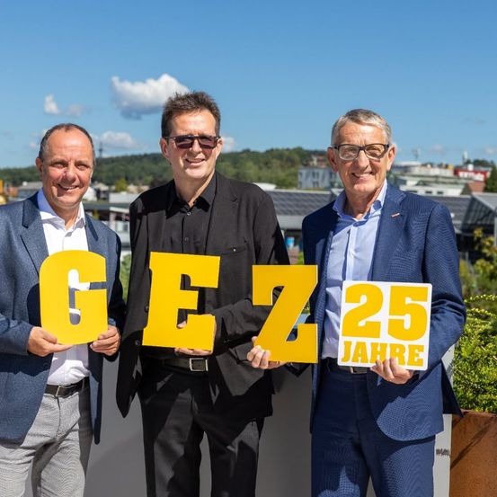 Drei Männer in Anzügen stehen zusammen und halten große gelbe Buchstaben, die 'GEZ 25 JAHRE' bilden. Sie lächeln und scheinen ein Jubiläum zu feiern. Hinter ihnen ist eine landschaftliche Ansicht einer Stadt mit Gebäuden und Bäumen.